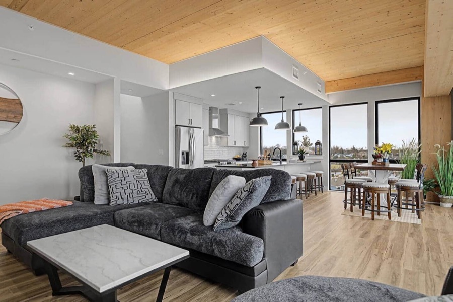Open-plan living room of Residence 601 with a plush charcoal sectional, wood-plank ceiling, and floor-to-ceiling windows over the Adelaide Pointe marina