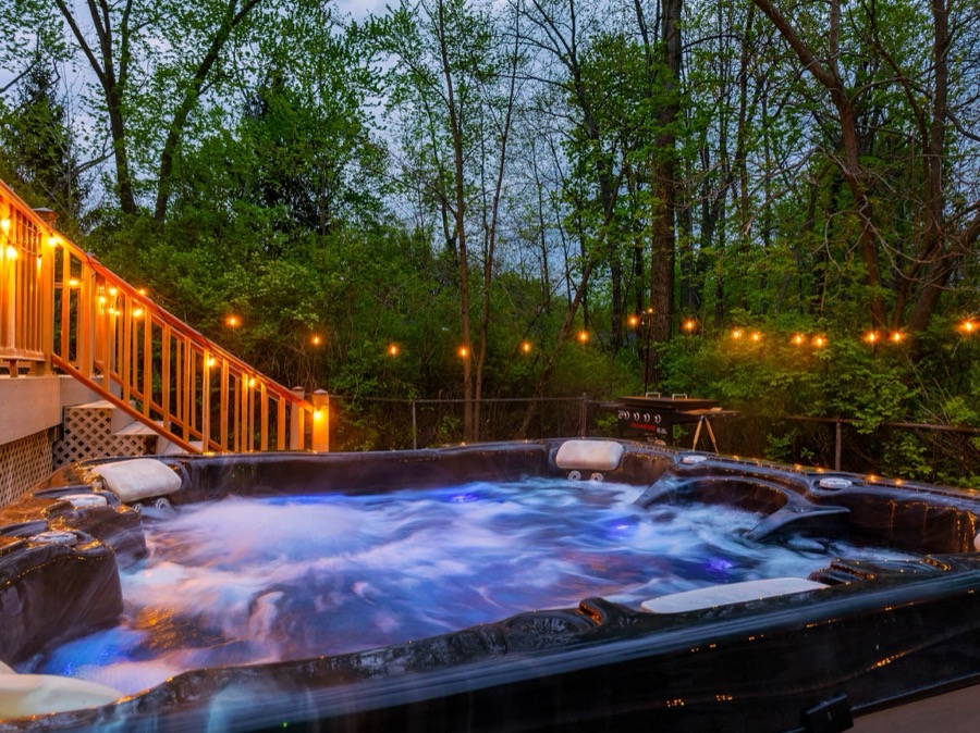Hot tub bubbling at dusk beneath the string-lit deck staircase, at the edge of the woods