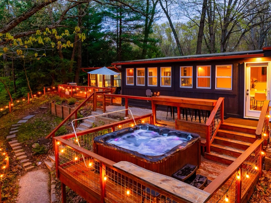 The hot tub bubbling on its own string-lit deck at twilight, with the cabin's window wall and bar-height deck seating glowing behind