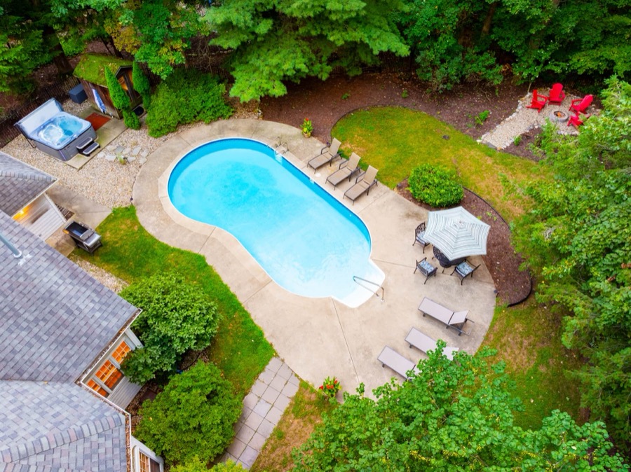 Aerial view of the backyard oasis: the kidney-shaped pool with loungers and umbrella table, hot tub and sauna on the gravel patio, and a red-chair fire-pit circle at the woods' edge