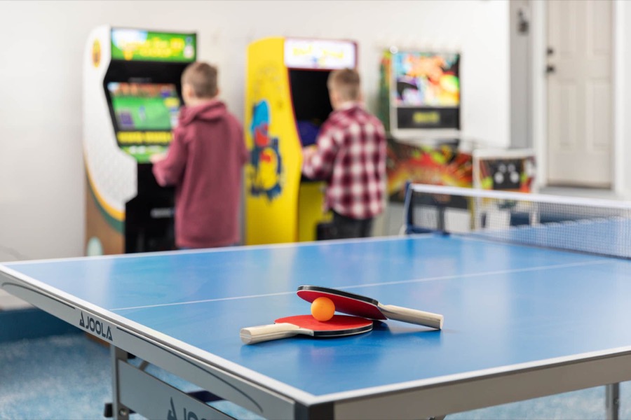 Kids at the arcade cabinets in the heated garage game room, ping pong paddles waiting on the table in front
