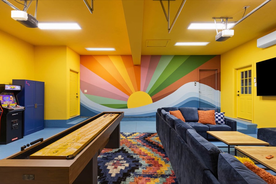 The garage game lounge: hand-painted sunset mural, shuffleboard table, arcade cabinet, and a huge navy sectional facing the TV